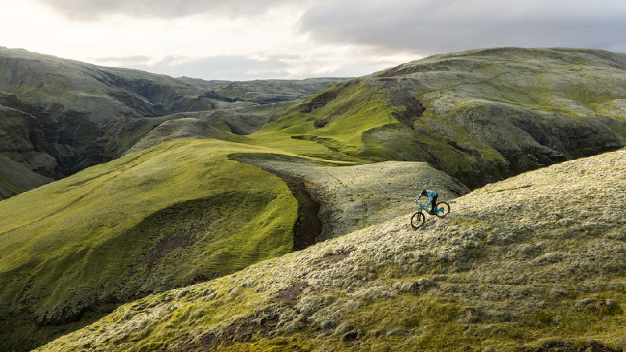 Un ciclista recorre un sendero angosto a través de colinas onduladas y verdes bajo un cielo nublado.