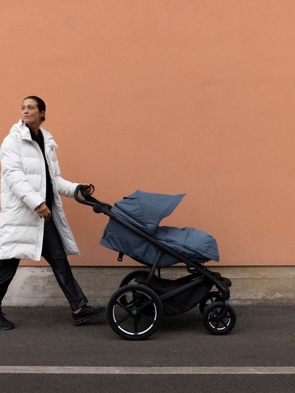 A woman walking with a dark blue Thule stroller with a footmuff in the same colour.