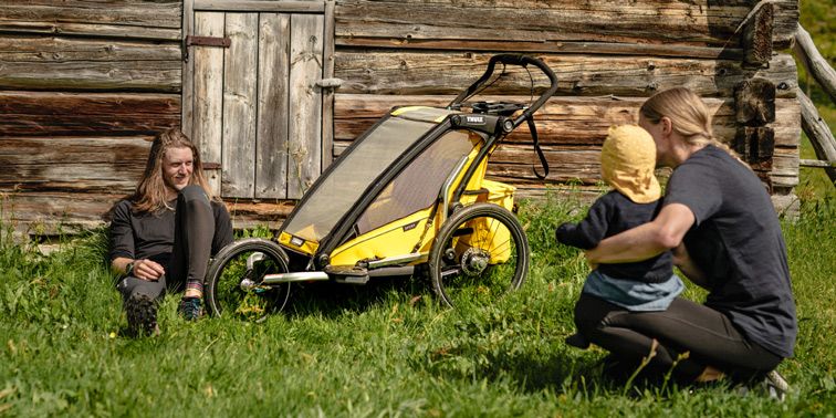 A man sits in the grass next to a yellow Thule Chariot Sport bike trailer with a Thule Orginzer sport accessory and a woman faces him with a baby on her lap.