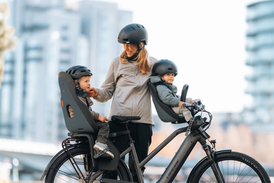 Adult standing with a bicycle fitted with Thule Yepp Nexxt 2 mini and Thule Yepp Nexxt 2 maxi seats, each carrying a child, on a city path with buildings in the background.