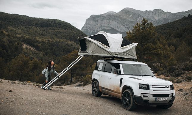 A vehicle is parked in the mountains with a Thule Approach rooftop tent that a woman is setting up. 