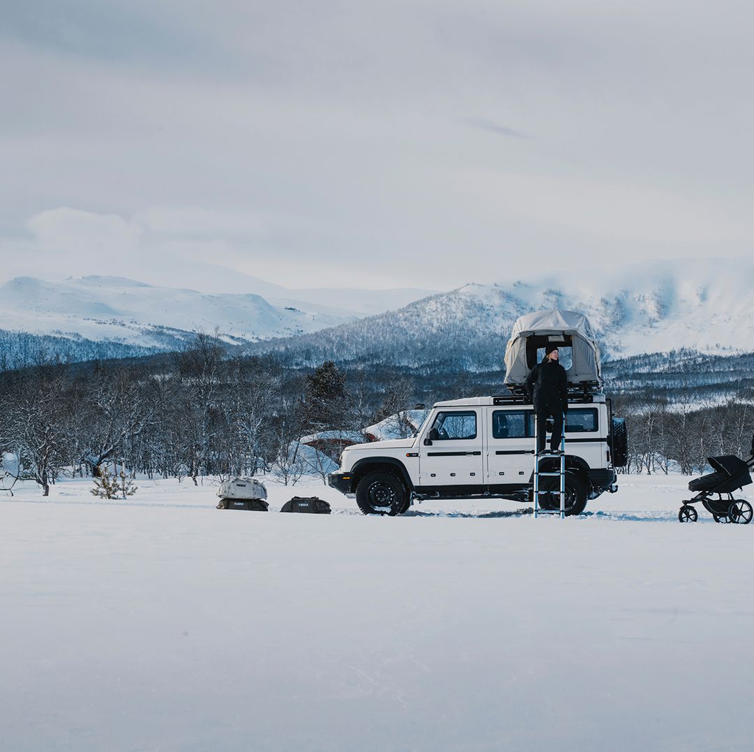 Eine Wintercamping-Szene mit einem weißen Geländewagen, der mit Ausrüstung von Thule ausgestattet ist, einschließlich eines Dachzeltes und anderer Outdoor-Utensilien, umgeben von verschneiten Bergen.
