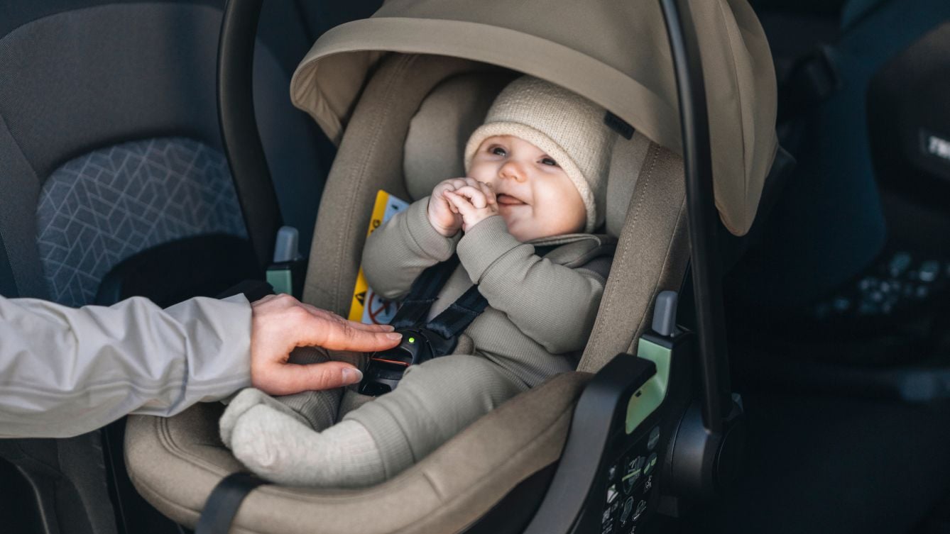 Baby sitting in a rear-facing Thule Maple Connect infant car seat while an adult fastens the e-Buckle inside a vehicle.