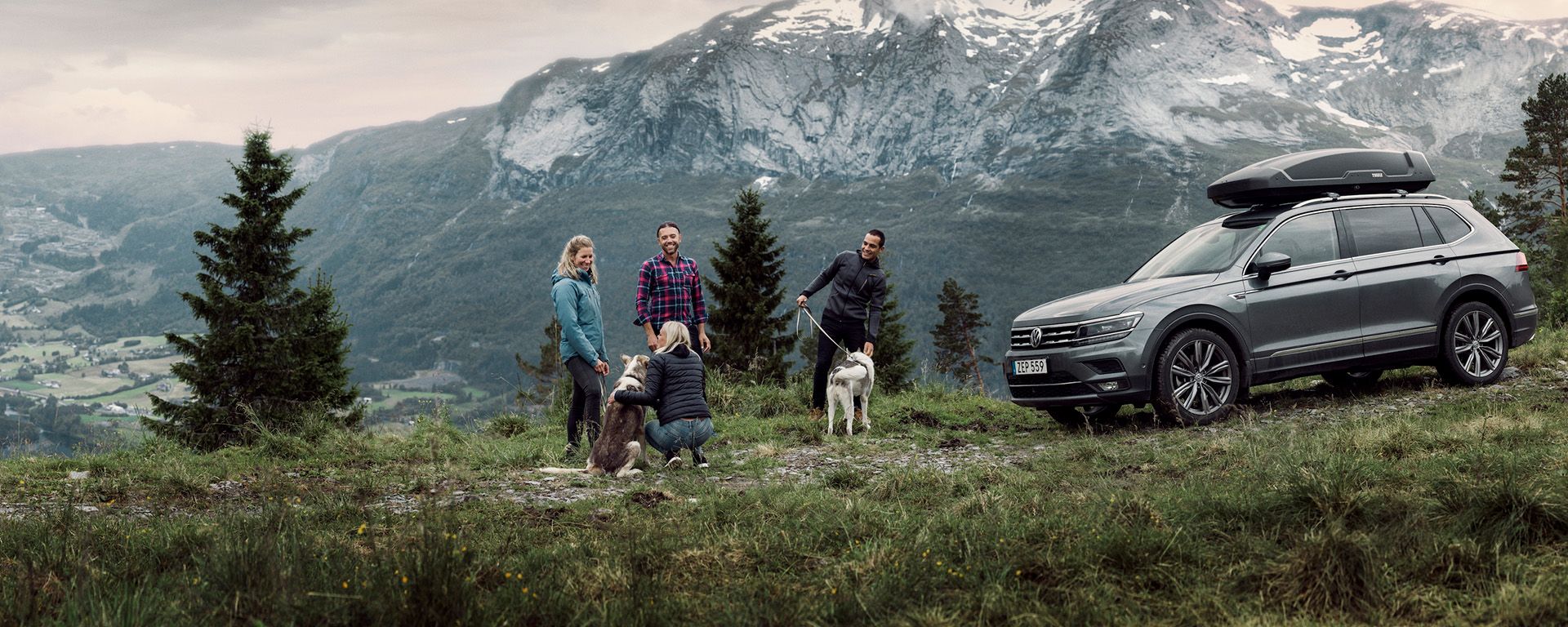 Drie vrienden staan met hun hond bij een auto met een dakkoffer in een landschap met besneeuwde bergen. 