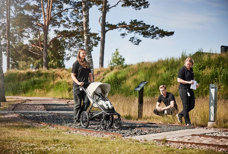 Une femme marche sur un terrain rocheux avec une poussette Thule Urban Glide à 4 roues pour tester sa stabilité, tandis que deux autres l'observent de près.