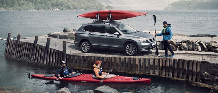 A man stands at the dock next to a car with a kayak rack, and a woman in a kayak in the water talks to him.