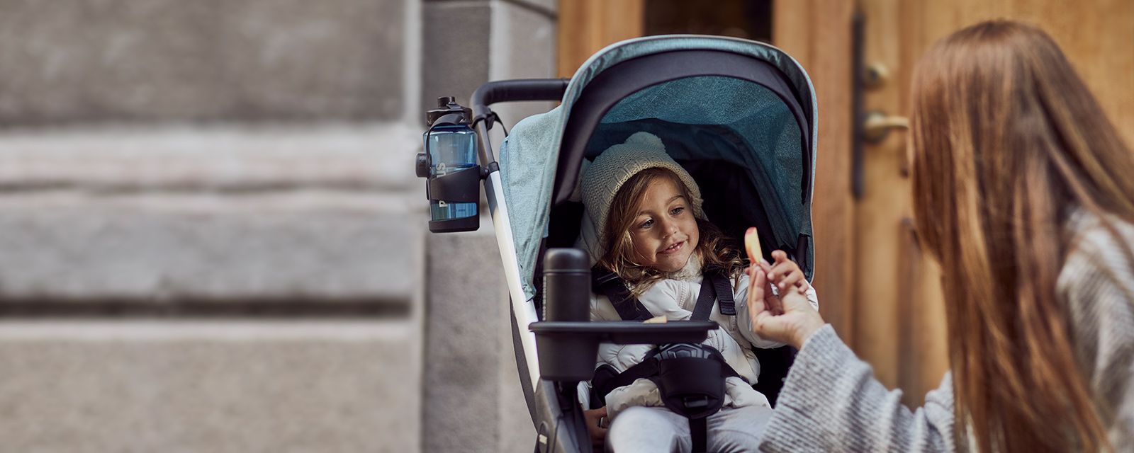 A woman hands a piece of apple to a child sitting in a thule stroller.
