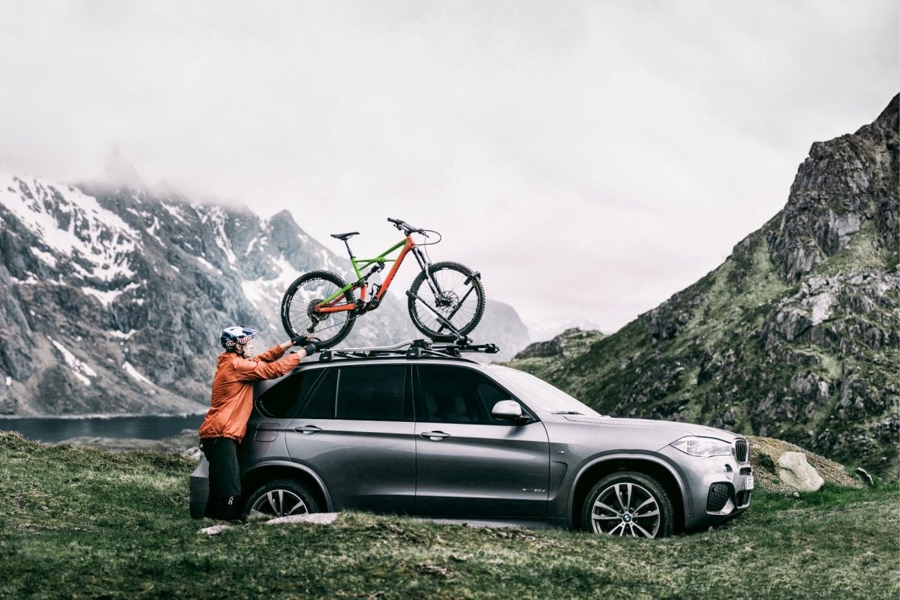 Un hombre carga su bicicleta en un portabicicletas con techo Thule con montañas y un lago al fondo.