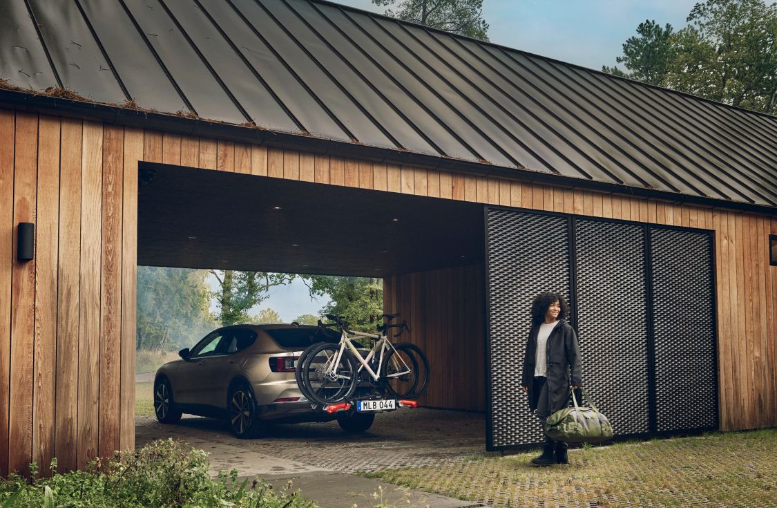 Um automóvel está estacionado numa garagem com um suporte de bicicleta Thule Epos montado na bola de reboque. Uma mulher caminha por trás do automóvel com um saco Thule Chasm verde na mão. 