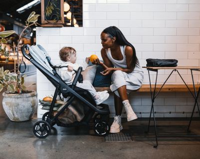 A woman sits on a bench holding an orange out to a baby in a Thule stroller.