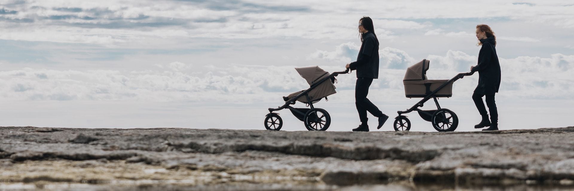 Two people walk along a rocky coastline with Thule Urban Glide strollers, showcasing their all-terrain design for outdoor adventures.