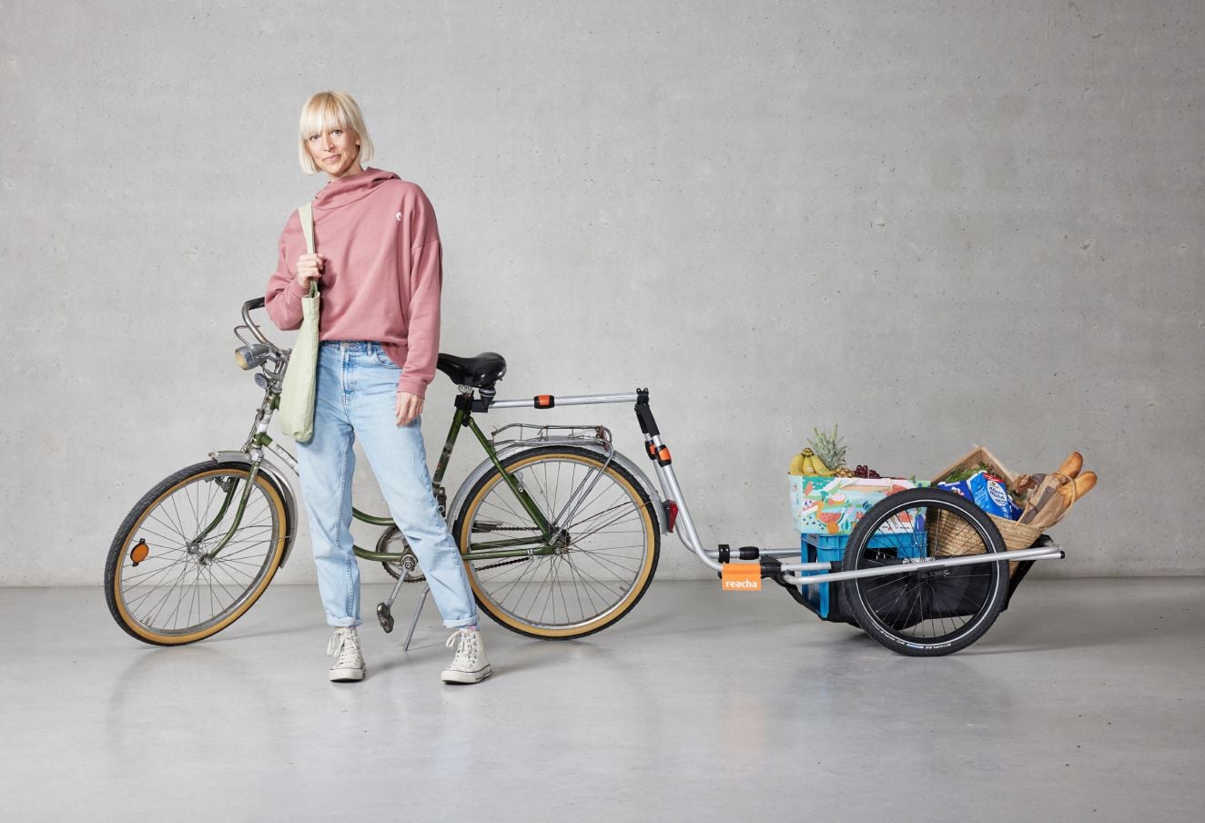 한 여성이 자전거에 부착된 Reacha City 자전거 트레일러와 함께 식료품과 기타 물건을 들고 스튜디오에 서 있습니다.
