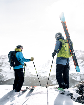 На горе стоят два человека с лыжами и лыжными рюкзаками.