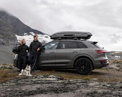 Tmavé SUV se střešní krabicí stojí na skalnatém svahu hor, zatímco vedle vozidla stojí dva lidé s venkovním vybavením a psem sedícím u jejich nohou. Místo obklopují sněhové plochy a strmé hory pod zataženou oblohou.