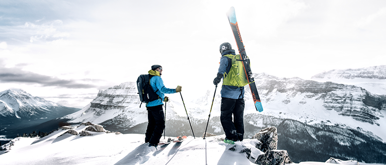 Two people stand on a mountain with skis and ski backpacks.