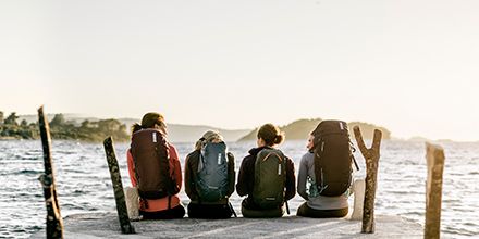 Four hikers sit on a boardwalk overlooking a lake at sunset, each carrying hiking packs.