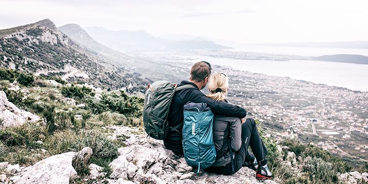 A woman and man look out over mountains and a city below, wearing Thule hiking backpacks.