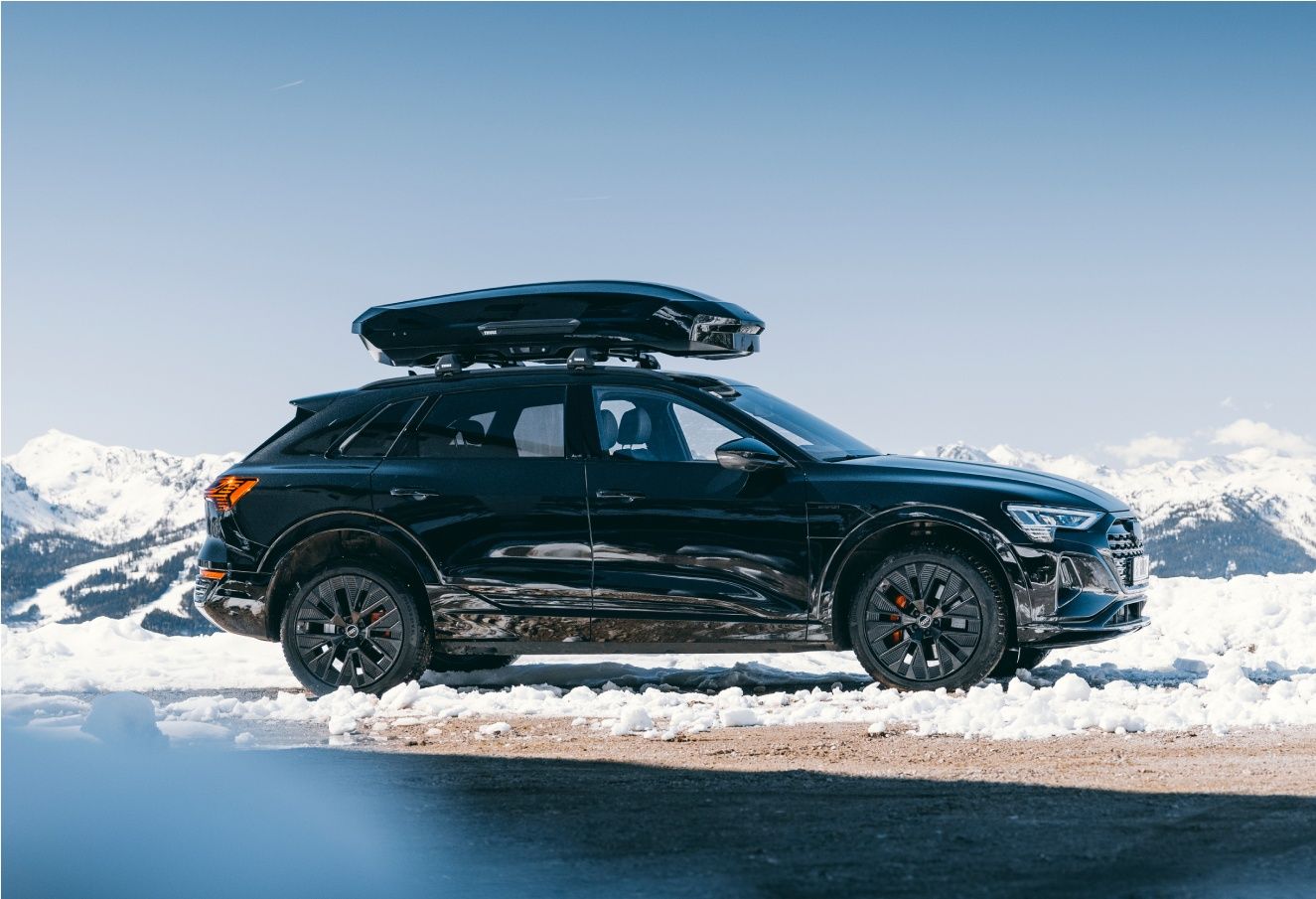A black SUV with a black Thule roofbox on top is parked in the snow with mountains in the background.