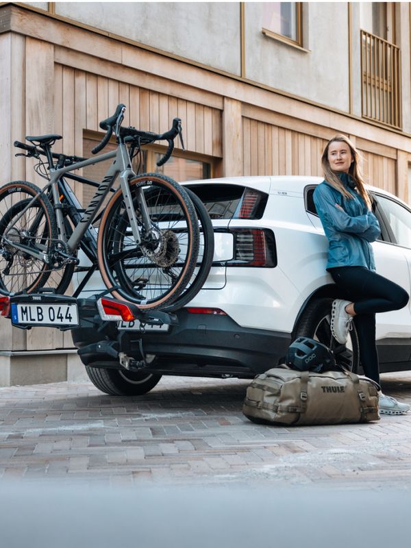 一名女子靠在車邊，腳邊放著一個 Thule Chasm 包款，車後方裝有 Thule Outpace 自行車架，穩固地載著兩輛自行車。