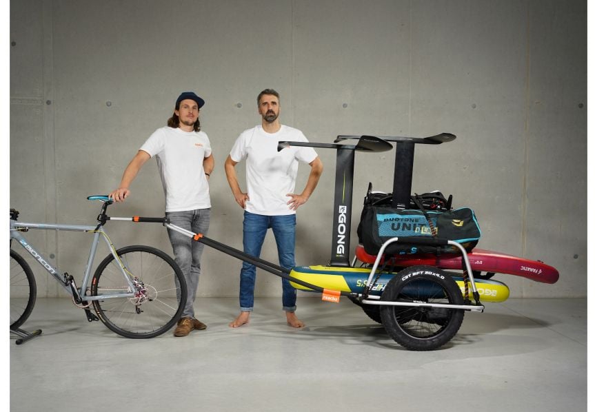 To menn poserer i et studio med en sykkel med Reacha Sport Beach-sykkelvogn med surfebrett og utendørsutstyr.