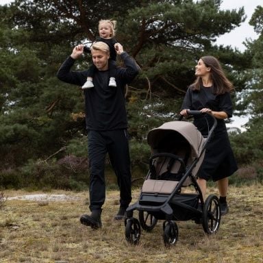 Muž s batoletem sedícím na ramenou kráčí přírodou po boku ženy, která tlačí prázdný kočárek Thule Urban Glide.