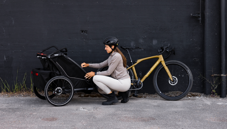 An adult kneels beside a bike trailer, adjusting it next to a parked bike.