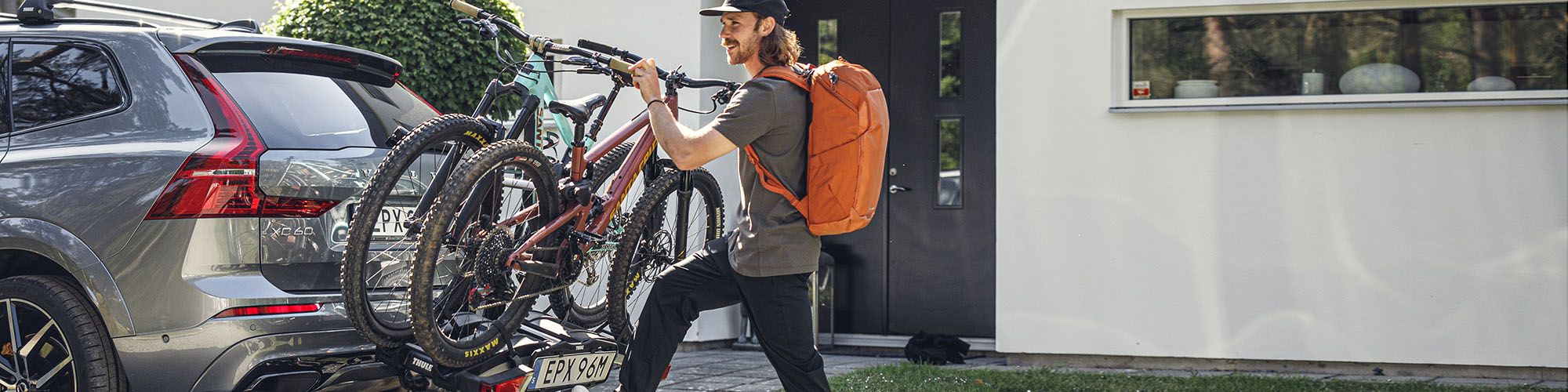 Man with an orange backpack lifts bikes onto hitch bike rack outside of a residential house.