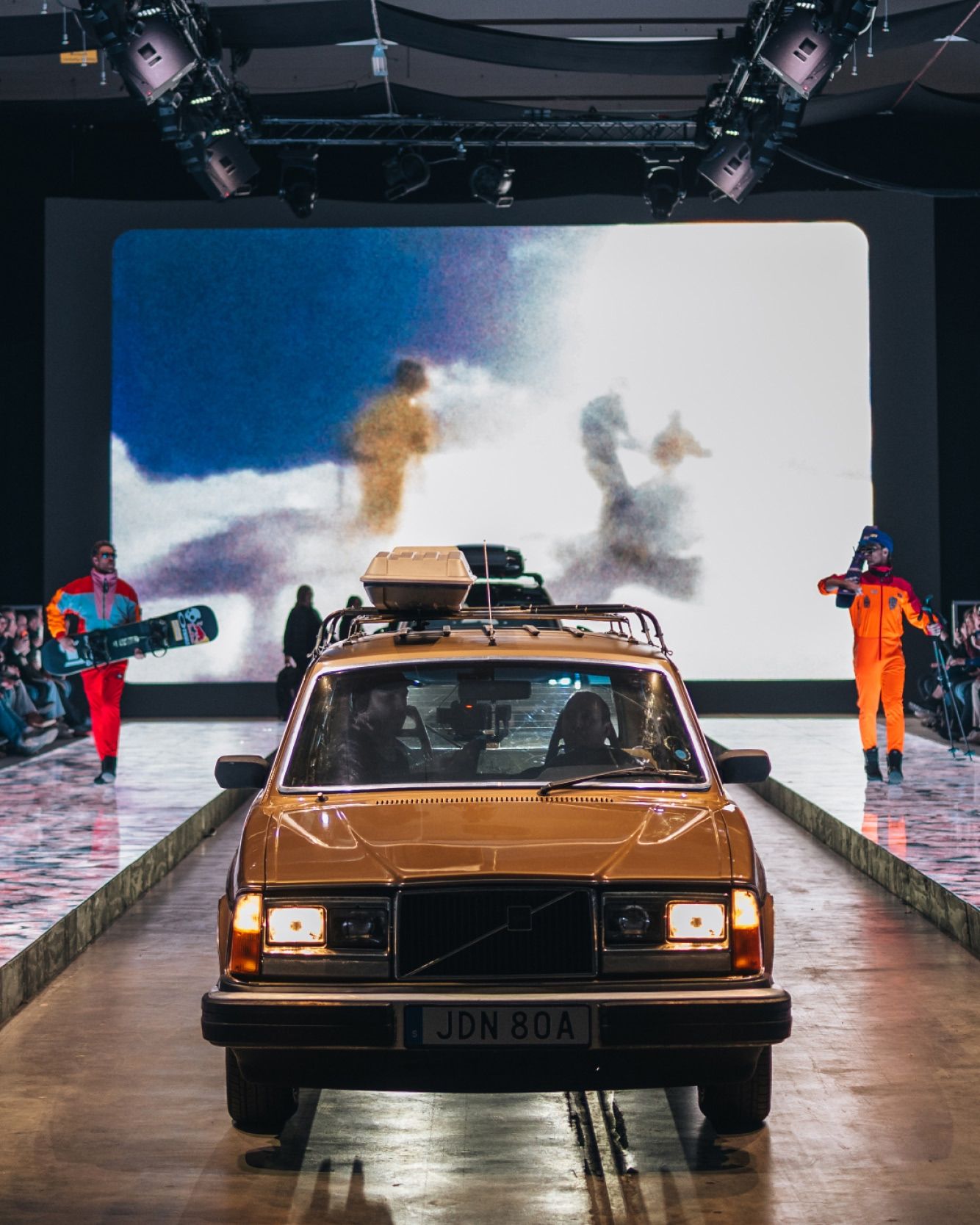 An older car with a roof box driving on a runway during a show, with performers holding snowboards in the background.