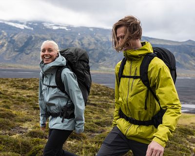 Two people hike through a field next to a mountain with hiking backpacks.