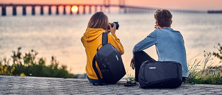Two people sit and watch the sun set behind a bridge, they both are carrying camera and lens backpacks.