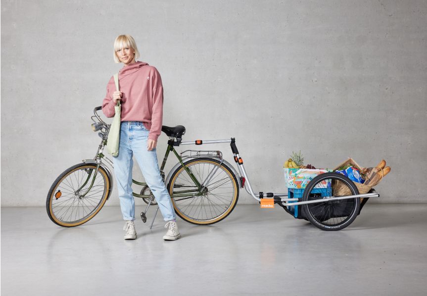 En kvinde står i et studie med en Reacha City-cykelanhænger, der er fastgjort til hendes cykel og bærer dagligvarer og andre genstande.
