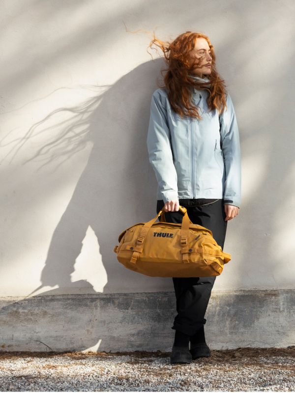 Femme debout contre un mur clair, tenant un sac fourre-tout Thule Chasm jaune, habillée pour l’aventure en plein air