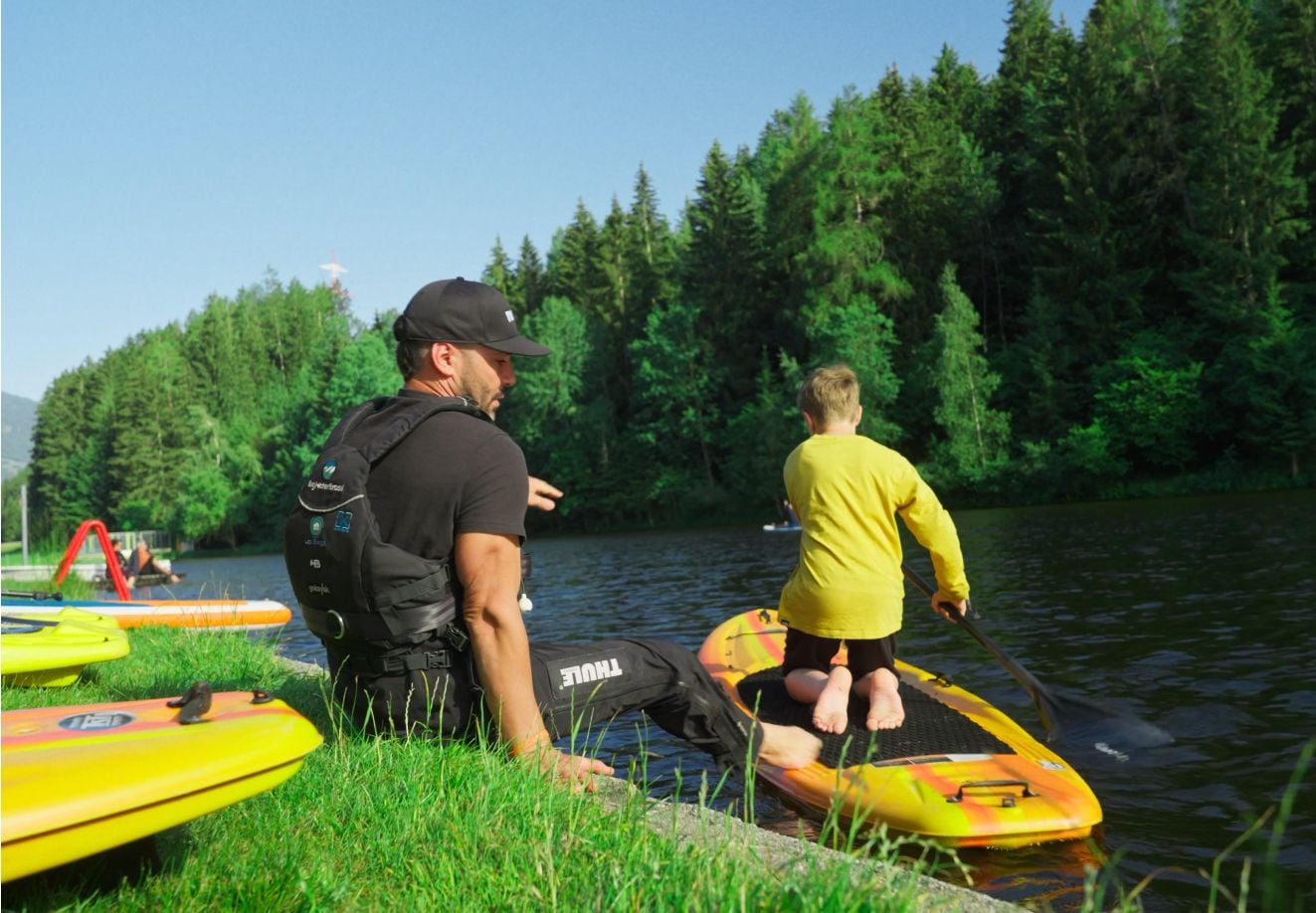 Dorosły siedzący na brzegu rzeki pomaga dziecku rozpocząć przygodę z deską SUP, delikatnie popychając malucha na wodę.