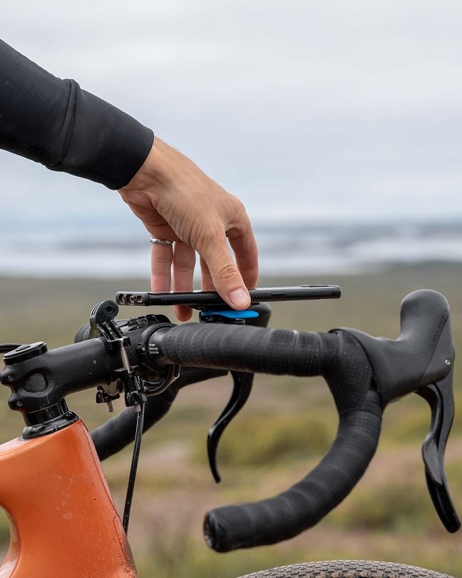 Close-up of a hand placing a smartphone onto a Quad Lock™ out front-mounted phone holder on a bicycle, with a blurred outdoor landscape in the background.