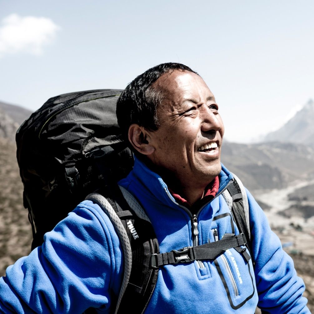 A close up image of Apa Sherpa smiling in the sunshine while wearing a backpack.