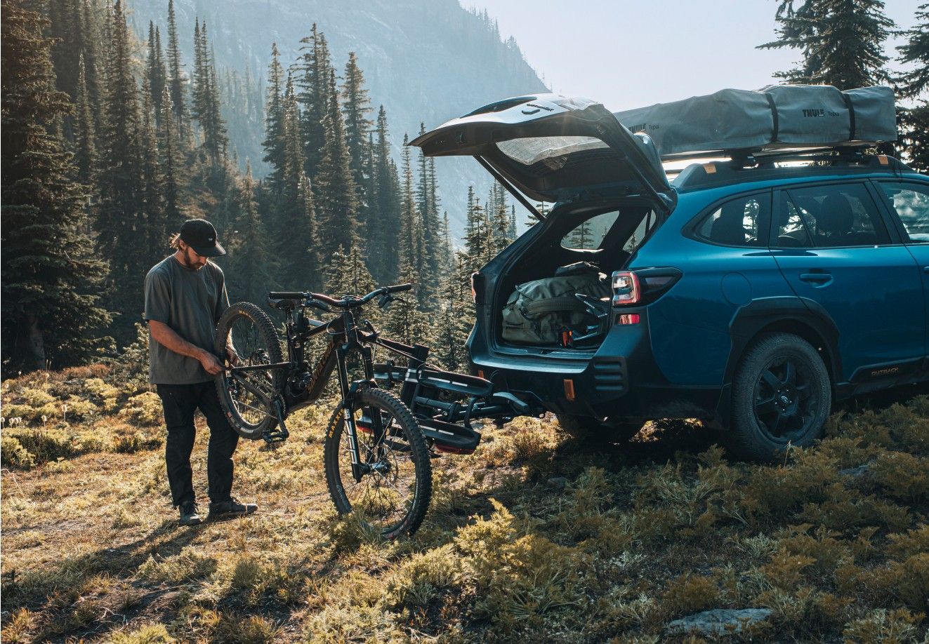 En mand reparerer sin mountainbike på en Thule-cykelholder monteret på anhængertræk bag på en blå Subaru Outback, parkeret i naturskønne bjergrige omgivelser med høje stedsegrønne træer og bjerge i baggrunden.