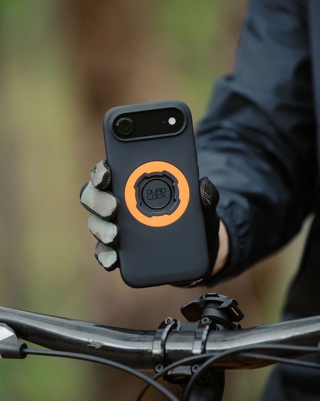 Close-up of a gloved hand holding a smartphone with a Quad Lock MAG™ phone cover mounting case above a bicycle handlebar.