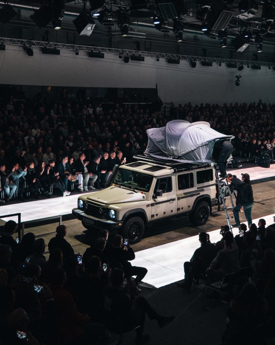 A vehicle with a rooftop tent displayed on a runway during a show, with an audience watching.