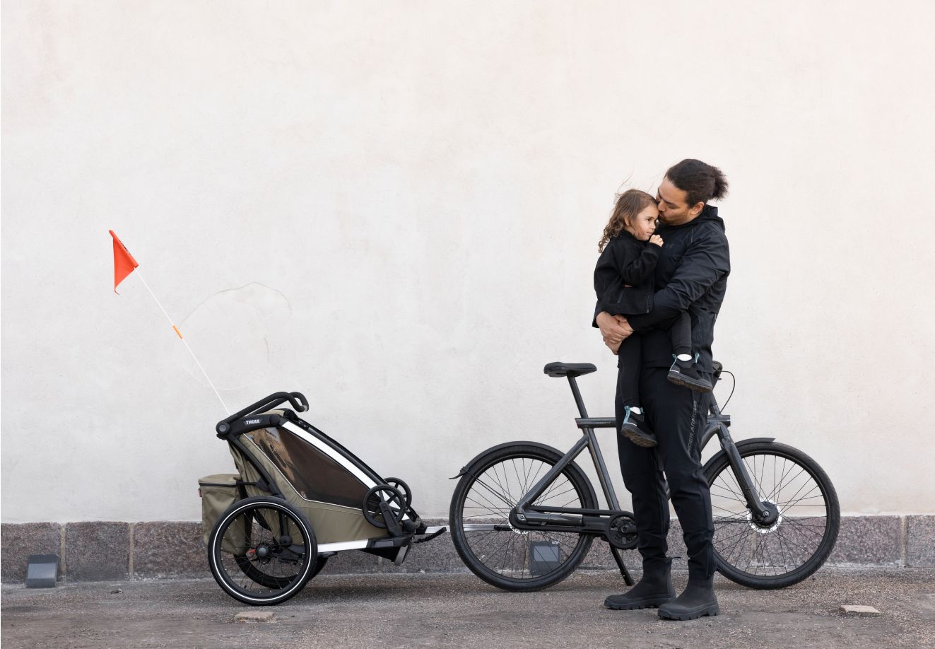 Un homme vêtu de noir se tient à côté d’un vélo auquel est fixée une remorque de vélo pour enfant. Il tient un petit enfant dans ses bras et ils semblent partager un moment de tendresse. La remorque de vélo est de couleur kaki et est pourvue d’un drapeau de sécurité orange.