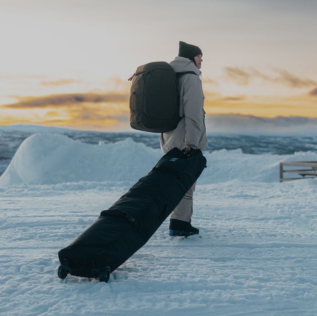Person carrying Thule ski bags through a snowy mountain landscape at sunset.