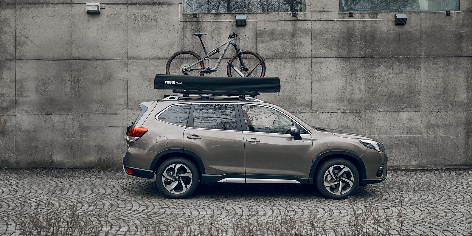 A vehicle parked next to a wall with a Thule Caprock roof platform, bike and rooftop tent on the roof.