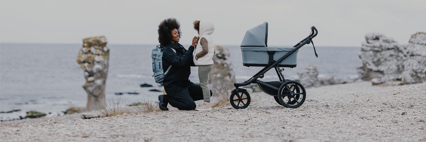 Una mujer que lleva una mochila Thule asegura un abrigo infantil en una playa junto a un carrito Thule. 