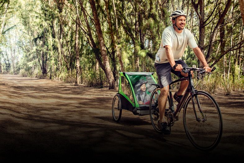 A man bikes on a bike path in the woods with his child in a Thule Cadence bike trailer for kids.